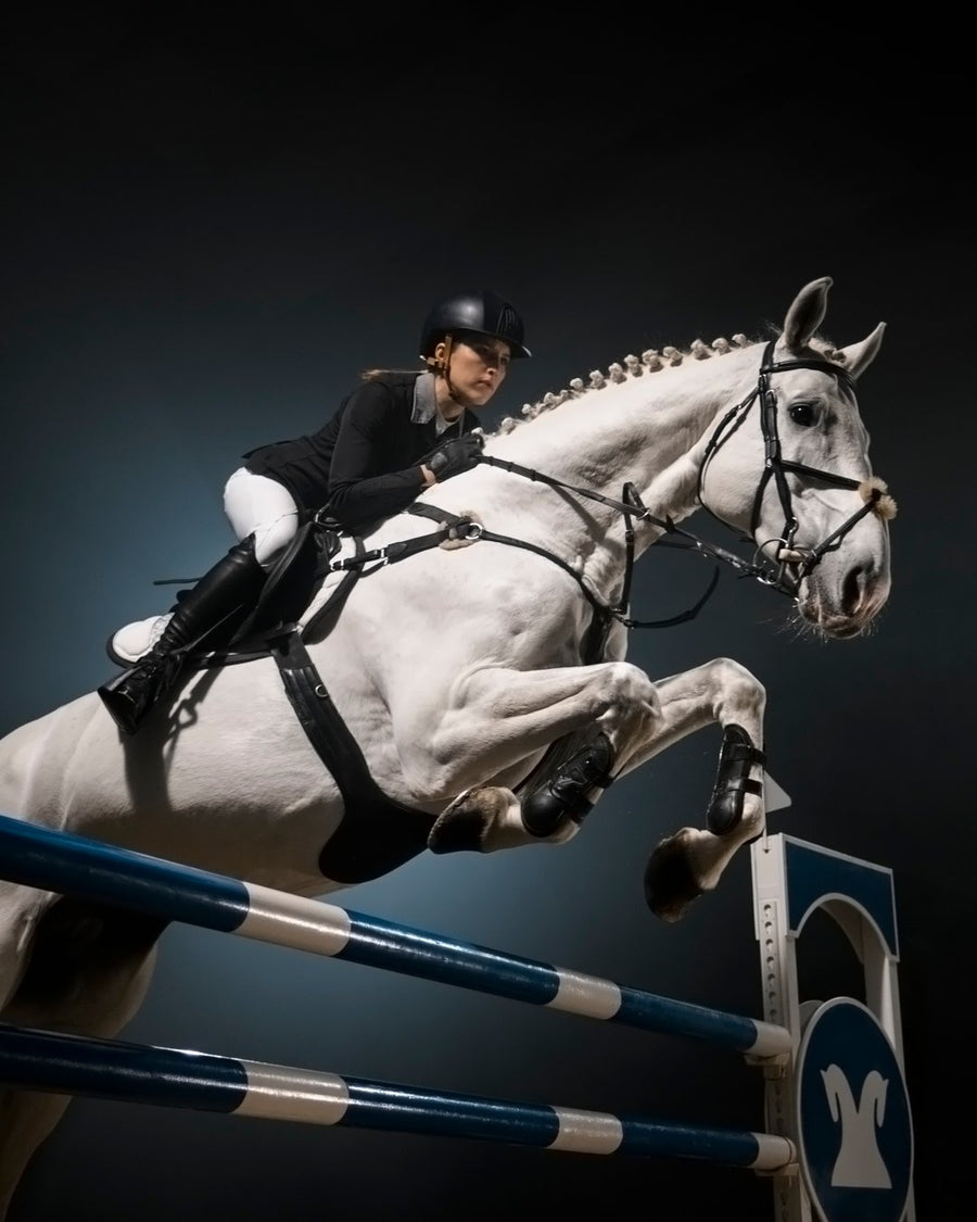 Show jumper in a black coat and helmet clearing a blue-and-white rail on a grey horse.