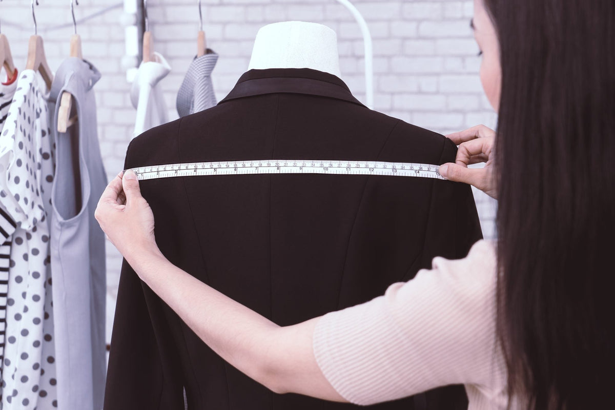 Tailor measuring across the shoulders of a dark jacket on a dress form with a tape measure
