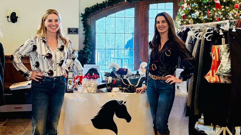 Two Tack and Tweed hosts stand beside their table of curated riding apparel at a holiday marketplace, with a Christmas tree behind them.