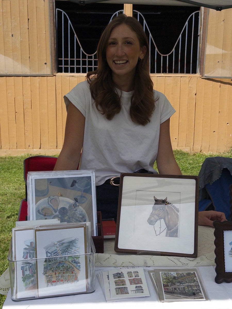 An artist smiling behind her table of equestrian prints and a framed pencil drawing of a horse, in front of the barn.