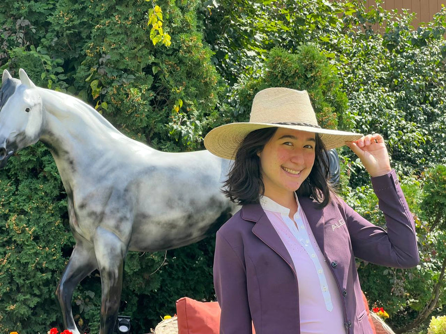 A woman in a straw hat and a plum show coat smiling beside a white horse statue in the garden.