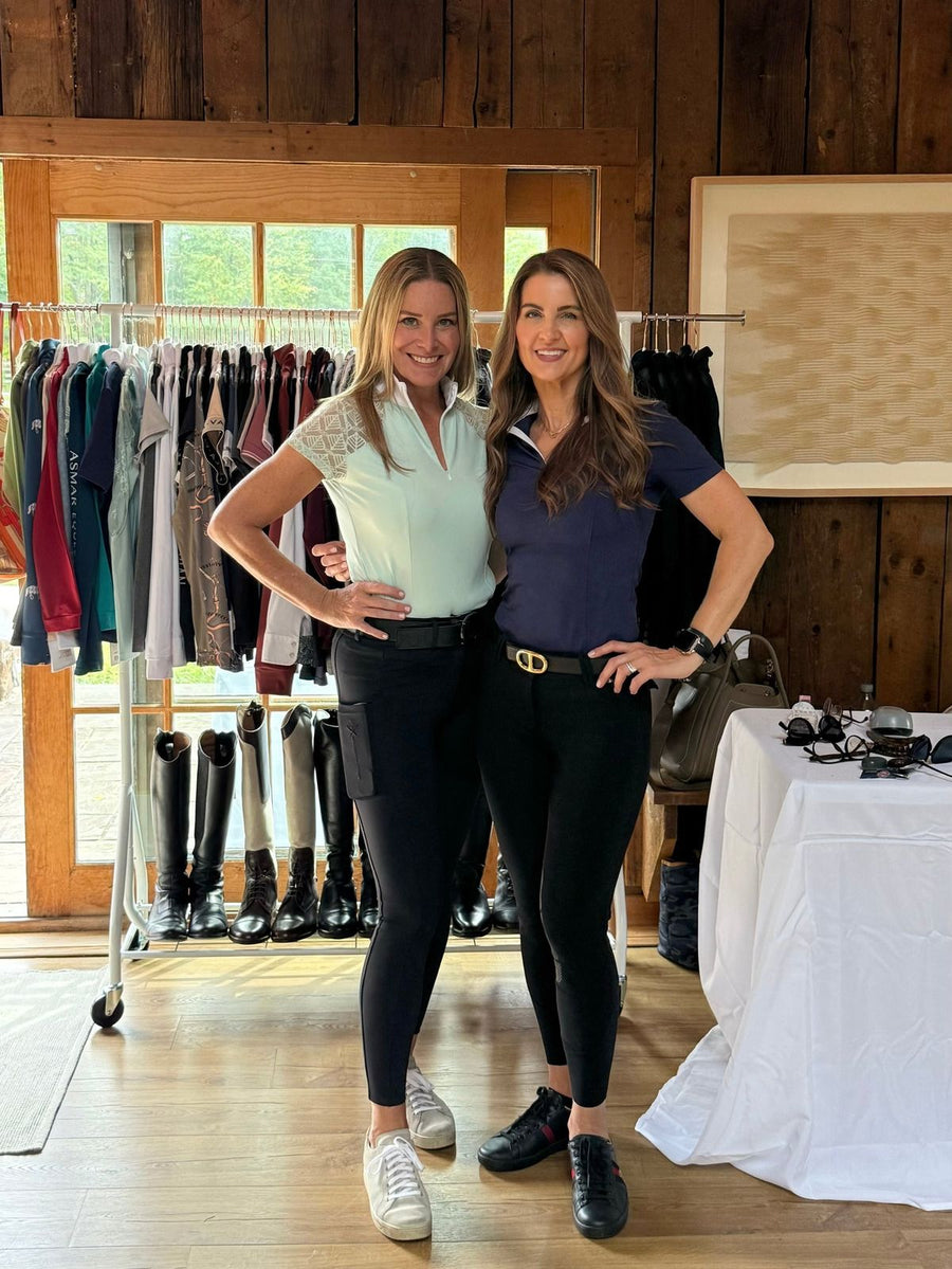 Two Tack and Tweed hosts in riding clothes in a barn lined with racks of apparel and tall boots.